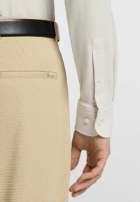 Beige textuur pants met een achterzak en een witte overhemd met lange mouwen en knoopmanchetten. Een zwarte riem maakt de outfit compleet.