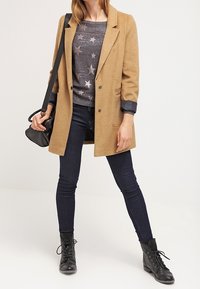 Brun ullblandad kappa med slagkrage och dubbelknäppning, tillsammans med en mörk tröja med silverstjärnor och mörka skinny jeans.