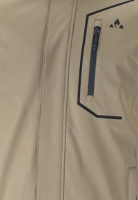 Beige jacka med en zippered bröstficka, svarta accenter och geometriska designelement. Mjuk, texturerad tyg med synliga sömmar.