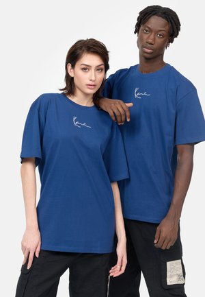 Blue cotton t-shirts with a relaxed fit and short sleeves, featuring a white signature logo on the chest. Black cargo pants are worn.