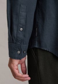 Donkerblauw geruit overhemd met een knoopmanchet, voorzien van zilveren knopen en een label met "Marc O'Polo" aan de zijkant. Glad textuur.