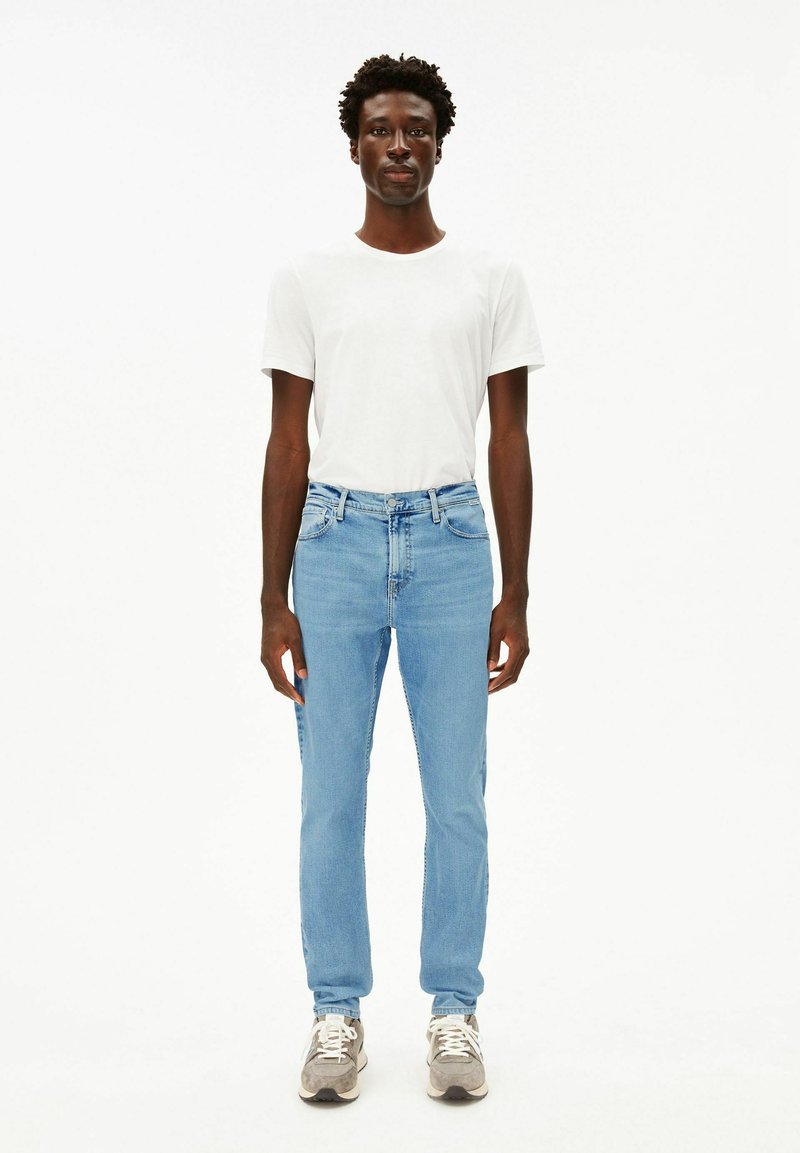 Hellblaue Jeans aus Denim mit figurbetontem Schnitt, kombiniert mit einem schlichten weißen T-Shirt. Das Modell trägt graue und weiße Sneakers.