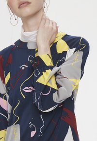 Monivärinen pitkähihainen paita, jossa on abstrakteja kuoseja punaisessa, keltaisessa, vaaleanpunaisessa ja harmaassa tummansinisellä taustalla; napitus yksityiskohtana hihoissa.