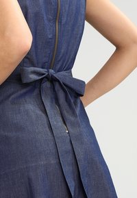 Denimové šaty s přiléhavým živůtkem, zadním zipem a mašlí v pase. Barva je tmavě modrá s jemnou strukturou.