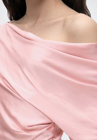 Kvinde iført en blød lyserød off-shoulder satin kjole med draperet stof, der viser hendes kraveben og bare skulder.
