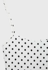 Haut blanc sans manches avec fines bretelles et un motif à pois noirs couvrant uniformément le tissu.