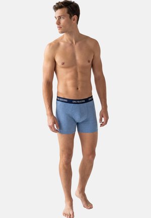 Bar overkrop mand iført blå boxer briefs med navy taljebånd mærket "ONLY & SONS," stående barfodet og kiggende mod venstre på en ensfarvet baggrund.