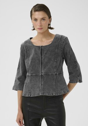 Žena s krátkymi hnedými vlasmi, ktorá nosí perlové náušnice, tmavosivú priliehavú džínsovú bundu s tromištvrťovými rukávmi a čierne nohavice na bielom pozadí.