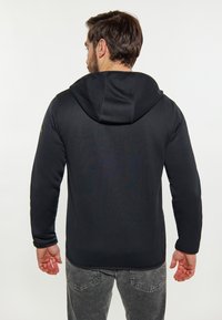 Felpa nera con texture a coste, cappuccio con cordino, maniche aderenti e orlo dritto, abbinata a jeans grigi in un contesto informale.