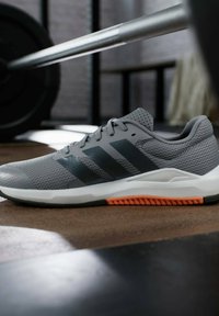 Zapatilla deportiva gris con parte superior de malla, que presenta tres franjas gris oscuro, una media suela blanca y un acento naranja en la suela. Diseño minimalista.