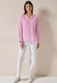 Blusa rosa chiaro a maniche lunghe con scollo a V e bottoni, abbinata a pantaloni bianchi dritti e sneakers beige. Texture liscia.