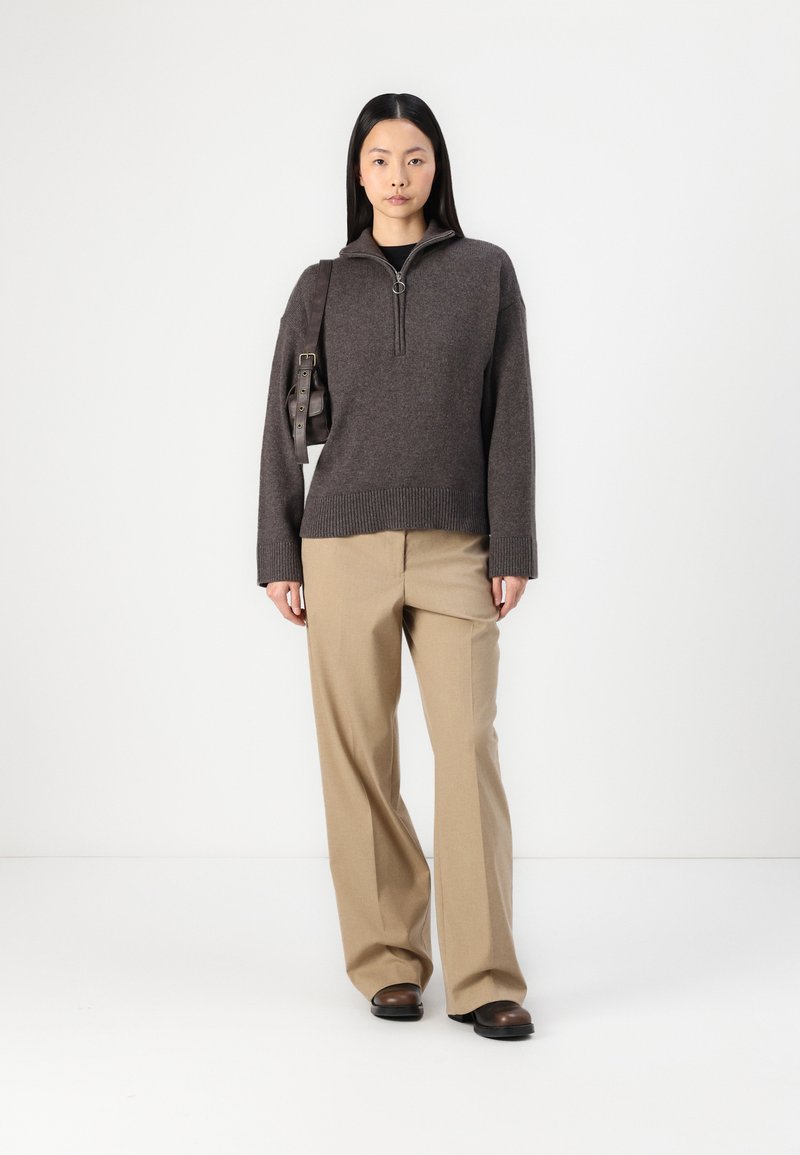 Ein charcoalfarbener Reißverschluss-Pullover mit geripptem Saum, kombiniert mit beigen weit geschnittenen Hosen und schwarzen Schuhen. Das Modell hält eine dunkelbraune Umhängetasche.