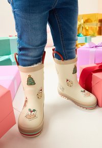 Cremfarbene Gummistiefel mit festlichen Aufklebern, darunter ein Weihnachtsbaum und Kekse. Mit roten Schlaufen als Griff und mehrfarbigen Sohlen ausgestattet.