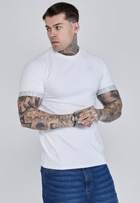 Fehér, rövidujjú póló, kerek nyakkivágással, szürke csíkkal az ujjakon és diszkrét márkázással a mellrészen. Puha, sima tapintású.