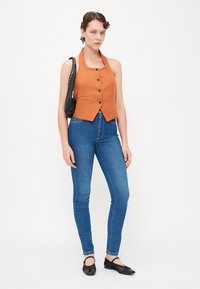 Rūsganbrūns, bez piedurknēm, pogājams vests ar pieguļošu dizainu, apvienots ar zilu džinsa skinny biksēm un melnām plakanu zoli kurpēm.