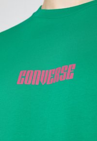 Zelené bavlněné tričko s jednoduchým designem, kulatým výstřihem a růžovou grafikou "CONVERSE" vytištěnou na přední části. Hladký povrch.