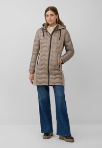 Abrigo tipo puffer beige con capucha, cierre de cremallera, patrón acolchado vertical, bolsillos laterales, combinado con jeans de mezclilla de pernera acampanada y botas negras de suela gruesa.