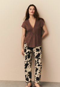 Bruine geplooide V-hals top in combinatie met beige broek met een zwart abstract vlekkenpatroon. Model draagt donkere sandalen; minimale achtergrond.