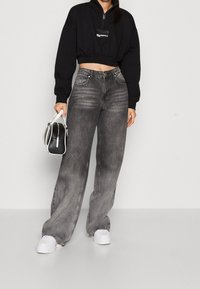 Svart croppad sweatshirt med dragkedja och logotyp, kombinerad med högmidjade, vida grå denimjeans. Modellen håller en liten vit handväska.