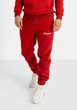 Des joggers en mélange de coton rouge présentent une taille élastique avec cordons de serrage et des chevilles élastiquées, affichant un logo blanc sur la jambe gauche.
