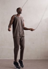 Haut sans manches marron et pantalon fuselé assorti portés par un homme, sautant à la corde, avec des chaussures de sport noires sur un fond neutre.