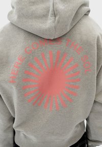 Sudadera gris con un gráfico de explosión solar impreso en rosa en la parte posterior, que presenta el texto "AQUÍ VIENE EL SOL" encima del diseño.
