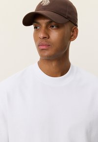 Καφέ καπέλο μπέιζμπολ με κεντημένο λογότυπο, φορεμένο με λευκό φούτερ με στρογγυλή λαιμόκοψη. Το καπέλο διαθέτει κυρτό γείσο και ρυθμιζόμενο λουράκι.