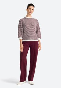Une femme se tient avec confiance, portant un pull rayé et un pantalon bordeaux, associés à des chaussures blanches, sur un fond gris clair.