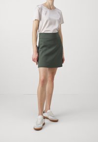Kvinna iklädd en ljusbeige kortärmad topp, mörkgrön knälång kjol, vita sneakers med gummisula och bär en vit axelremsväska.