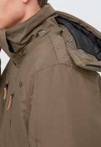 Ruda, oro sąlygoms atspari striukė su aukštu apykakle, reguliuojama kapišonu ir užtrauktukais užtraukiamomis kišenėmis; vidinė pamušalas juodas, o akcentai - odiniai.