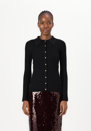 Schwarze gerippte langärmlige Strickjacke mit spitzem Kragen, die eine Reihe runder Knöpfe auf der Vorderseite aufweist. Trägt man über einem paillettenbesetzten burgunderfarbenen Rock.