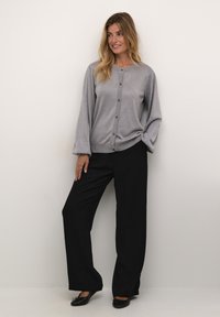 Ljust grå knäppt cardigan med långa ärmar, tillverkad av ett strukturerat metalliskt tyg, kombinerad med svarta vida byxor och platta skor.