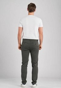 Hombre de pie de espaldas, vistiendo una camiseta blanca de manga corta, pantalones gris oscuro y zapatillas blancas, contra un fondo neutro.