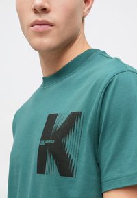 Tyrkysové bavlněné tričko s černým grafikou stylizovaného "K" s horizontálními čarami. Kulatý výstřih a krátké rukávy.