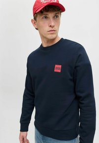 Marineblauer Sweatshirt mit geripptem Rundhalsausschnitt und -bündchen, ausgestattet mit einem roten quadratischen Logopatch auf der linken Brust. Getragen mit hellblauen Jeans.