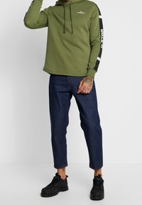 Olivgrön huvtröja med framficka och dragsko på huvan; svarta och vita accenter på ärmarna. Kombinerad med mörkblå tajta jeans och svarta sneakers.