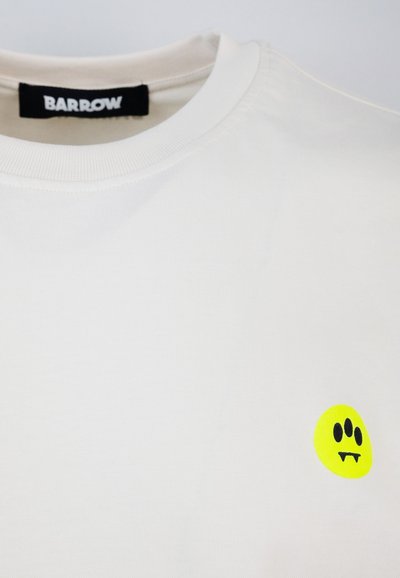 T-shirt in cotone bianco con scollo rotondo e una vivace grafica gialla di smiley con accenti neri sul petto. Etichetta del marchio visibile.