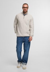 Hellbeige Pullover mit einem halben Reißverschlusskragen, über einem gemusterten Hemd getragen. Dazu blaue Jeans und beige Sneakers.