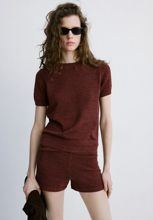 Kobieta o falowanych włosach, ubrana w czarne okulary przeciwsłoneczne, bordowy dzianinowy krótki top z krótkim rękawem i dopasowane szorty, trzymająca w jednej ręce ciemny element garderoby.