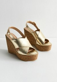 Sandales compensées métalliques dorées avec plateforme en liège, design à lanières croisées, bout ouvert et bride ajustable à la cheville. Finition lisse et silhouette épurée.