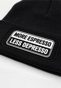 Black knit beanie with ribbed texture, featuring a white tag that reads "MORE ESPRESSO LESS DEPRESSO" on a rectangular patch.