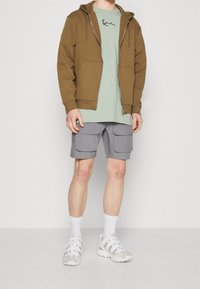 Hnědá mikina na zip, světle zelené tričko s černým logem, šedé cargo šortky s velkými kapsami a bílé sportovní boty.
