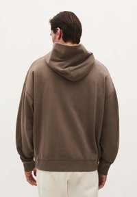 Brązowa, oversize bluza z dużym, luźnym kapturem, prążkowanymi mankietami i dołem. Miękka tekstura, minimalistyczny design. Noszona z jasnymi spodniami.