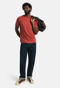Red crew-neck t-shirt, dark blue denim jeans, brown and beige sneakers with white laces. Model holding a navy bag over one shoulder.