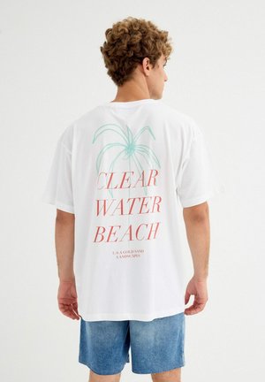 Hombre con cabello rizado que lleva una camiseta blanca extragrande con la inscripción "CLEAR WATER BEACH" y un gráfico de una palmera, combinado con pantalones cortos de mezclilla azul.