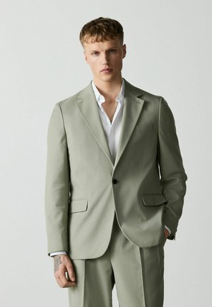 Young man with short blond hair wearing a light gray suit and white shirt, standing with one hand in pocket against plain background.