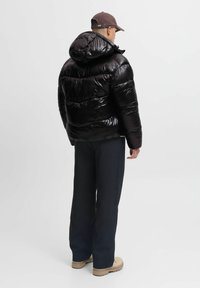 Uomo che indossa un giubbotto imbottito nero lucido con cappuccio, pantaloni scuri, stivali beige e un cappellino da baseball marrone, in piedi e girato di spalle su uno sfondo bianco.