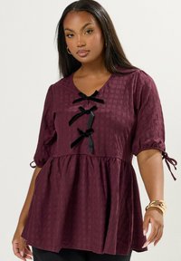 Donna che indossa una blusa di prugna texturizzata con tre fiocchi neri lungo il davanti e maniche a metà legate alle estremità, accessoriate con braccialetti dorati.
