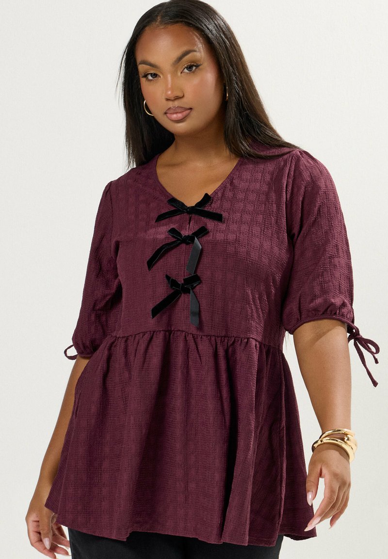 Donna che indossa una blusa di prugna texturizzata con tre fiocchi neri lungo il davanti e maniche a metà legate alle estremità, accessoriate con braccialetti dorati.