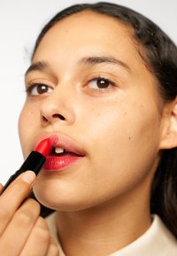 Rode lippenstift met een gladde textuur wordt op de lippen aangebracht. De lippenstift heeft een zwarte behuizing, en het model heeft een gemiddelde huidtint met zichtbare sproetjes.